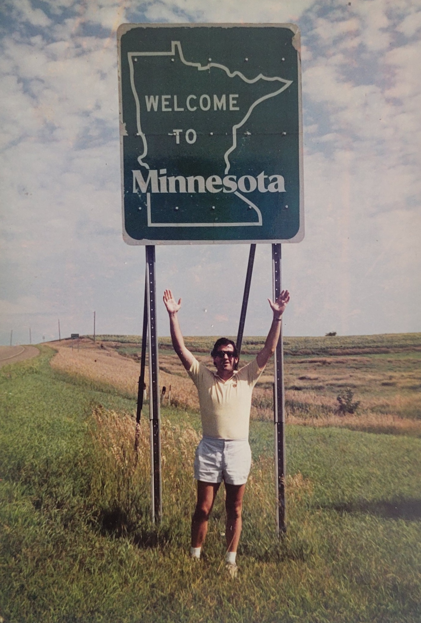 A young John Millea raises his arms to the sky while standing under a "Welcome to Minnesota" highway sign.