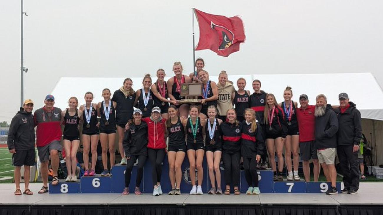 Alexandria Area Girls Track and Field team on MSHSL podium with Cardinals flag and first-place trophy
