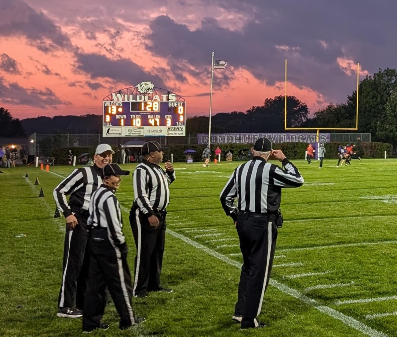 Officials talk and laugh under a colorful, cloudy sky during halftime of a high school football game.